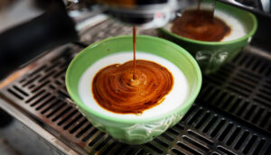 A cafe au lait being brewed: heated milk in a bowl, with espresso brewing into it.