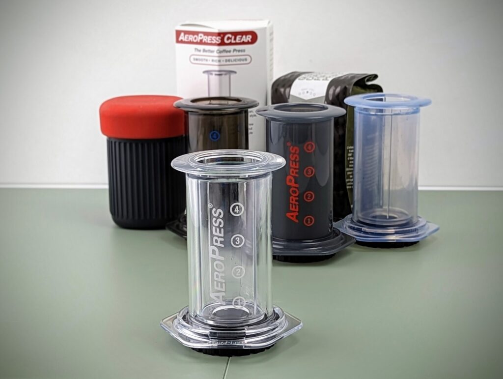 The current AeroPress Clear, photographed with some older versions of the AeroPress, including (left to right) AeroPress Go, Aeropress 2011 model, Aeropress 2022 model, and an AeroPress Prototype.
